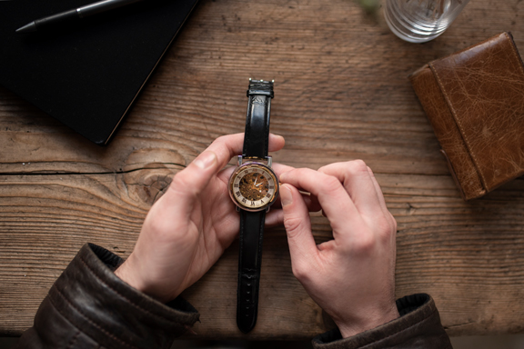 Person stellt eine Armbanduhr mit schwarzem Lederarmband auf einem Holztisch ein.
