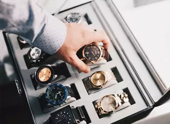 Hand greift nach einer Armbanduhr in einer Uhrenbox mit verschiedenen Modellen – Symbolfoto für Tipps zum Uhrenverkauf